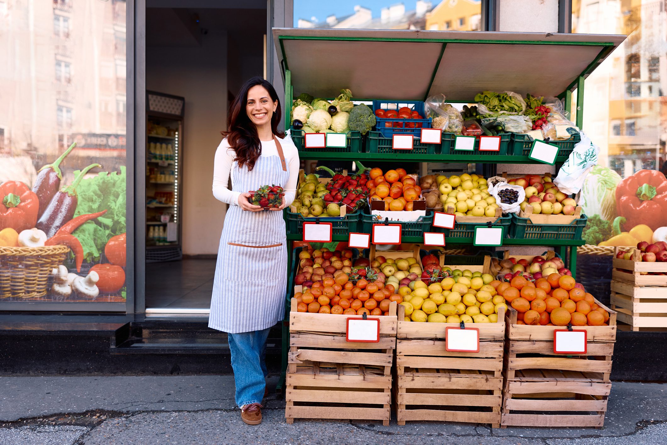 Smiling woman wearing an apron holding a basket of fresh strawberries, standing in front of an outdoor produce display at a small business grocery store entrance, offering healthy food to customers